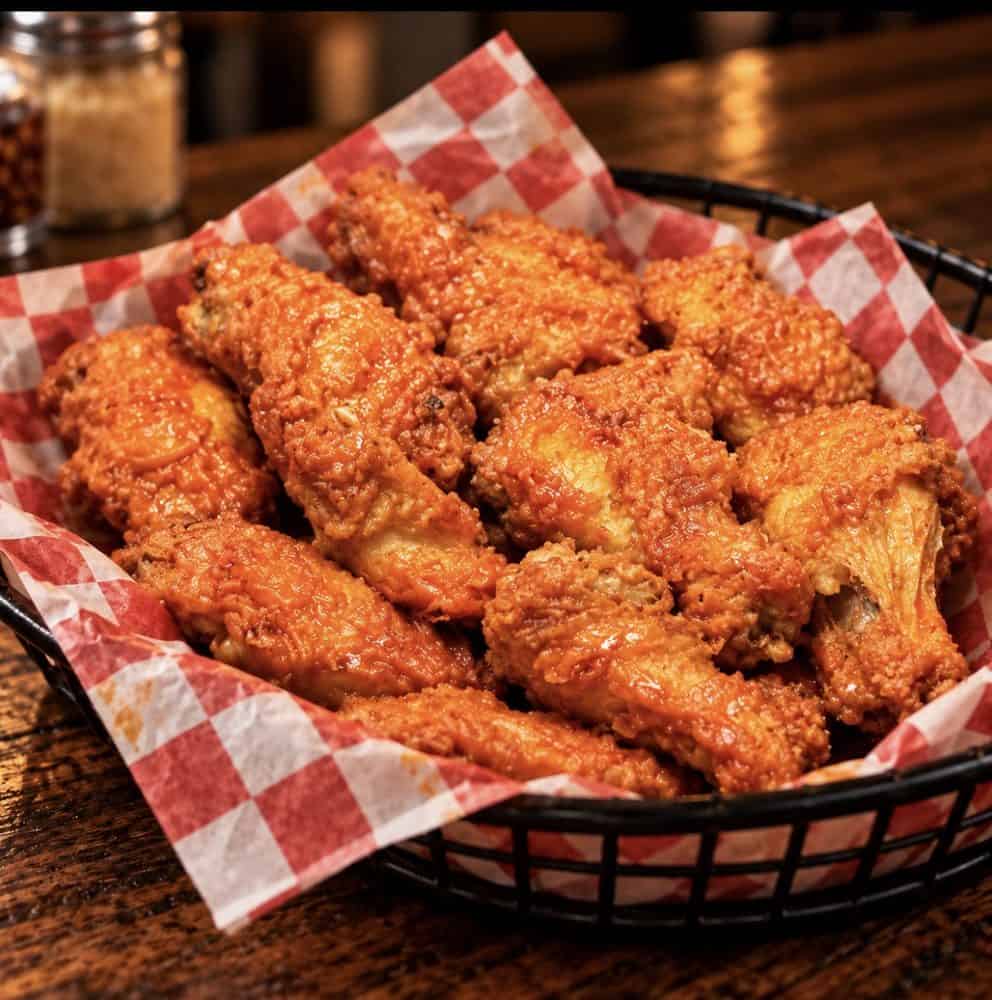 Buffalo chicken wings coated in orange-red sauce in a basket lined with red-and-white checkered paper.